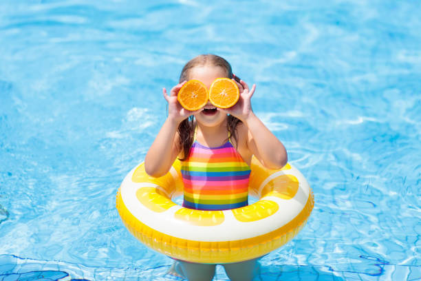 Little girl in swimming pool with inflatable toy ring eating orange. Kids swim on summer vacation. Tropical fruit and healthy snack. Swim aids for child.  Kid on colorful float. Beach and water fun.